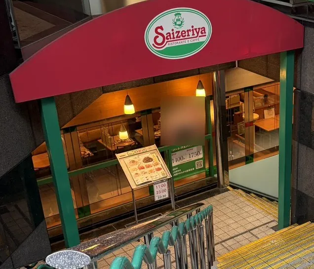 萨莉亚(Saizeriya)门店入口。红色屋顶上有绿色椭圆形的 Saizeriya 招牌,入口前立着菜单架和营业时间牌。