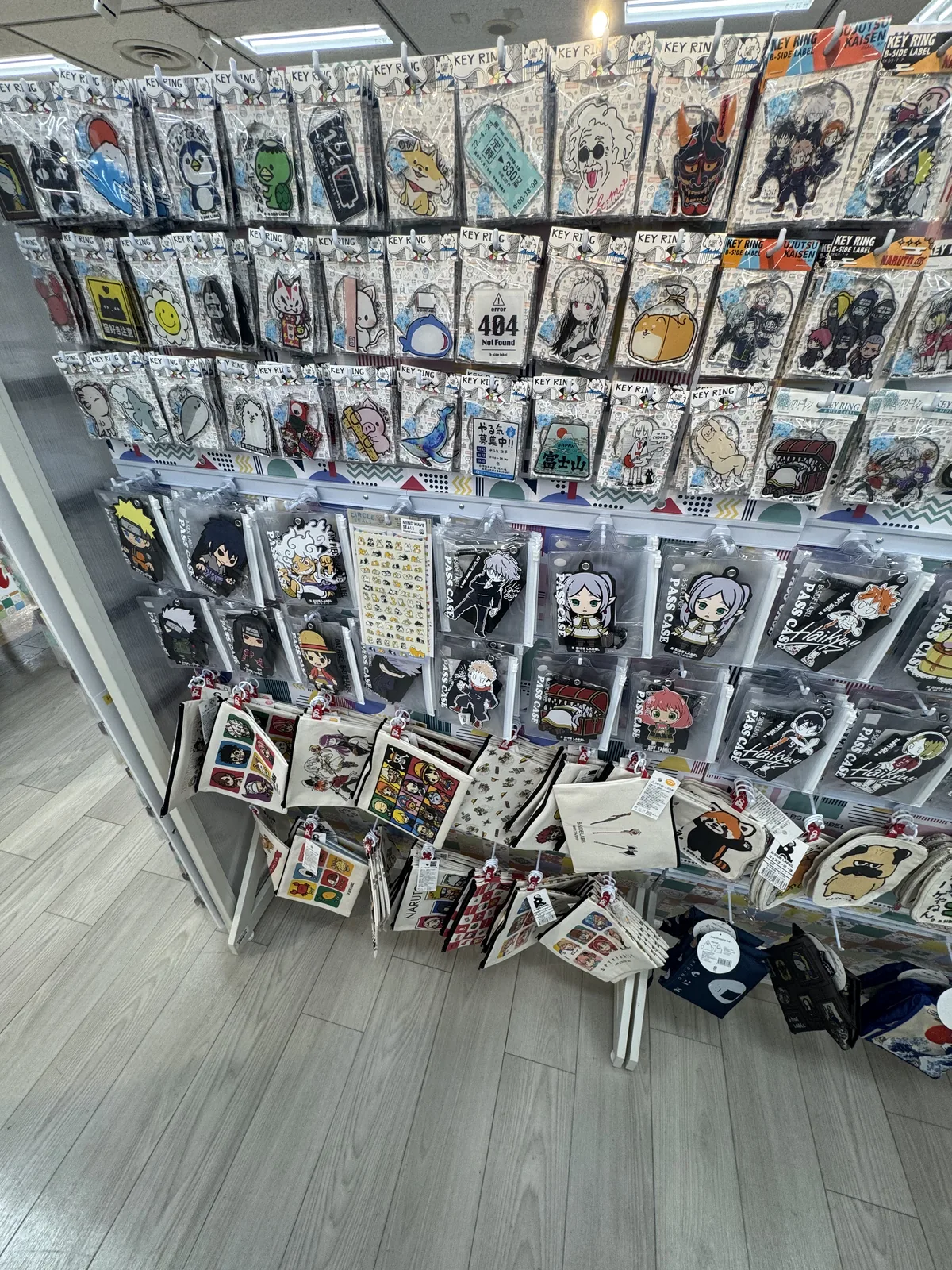 Portachiavi, tra cui quelli di personaggi degli anime, e portatessere appesi a uno scaffale; più in basso sono appese delle borse di tela.