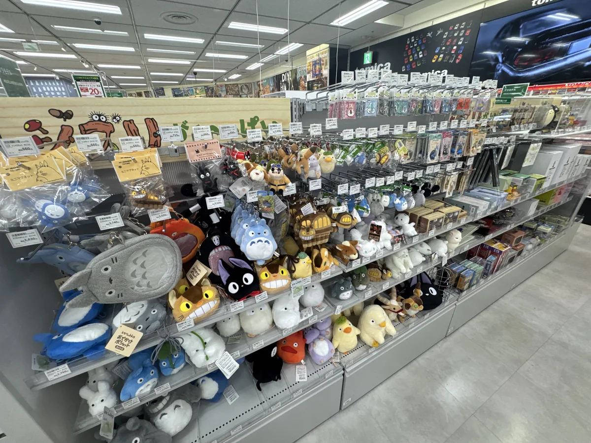 Peluche dei personaggi delle opere dello Studio Ghibli allineati su uno scaffale. Sullo scaffale in fondo sono appesi dei portachiavi.