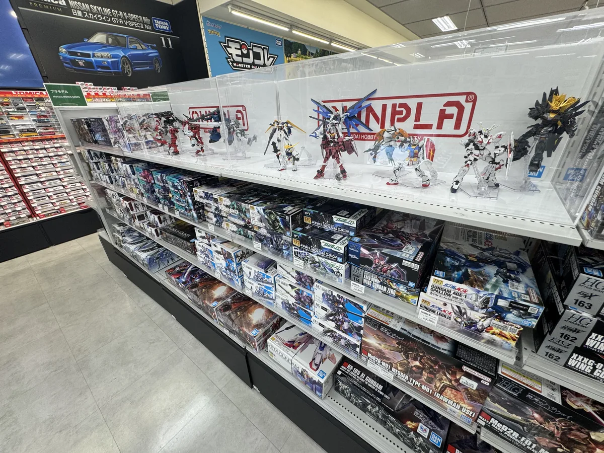 Scatole di modellini in plastica di Gundam allineate sugli scaffali; in una vetrina trasparente in alto sono esposti modelli già montati.