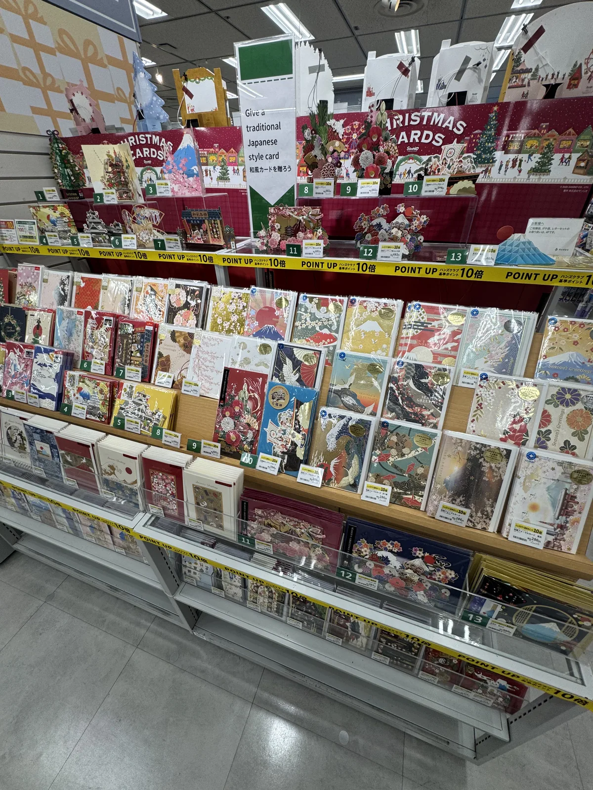 Uno scaffale di biglietti d'auguri. Ci sono biglietti con motivi giapponesi come il monte Fuji, le gru e i fiori, e un cartellino con la scritta “Give a traditional Japanese style card”.