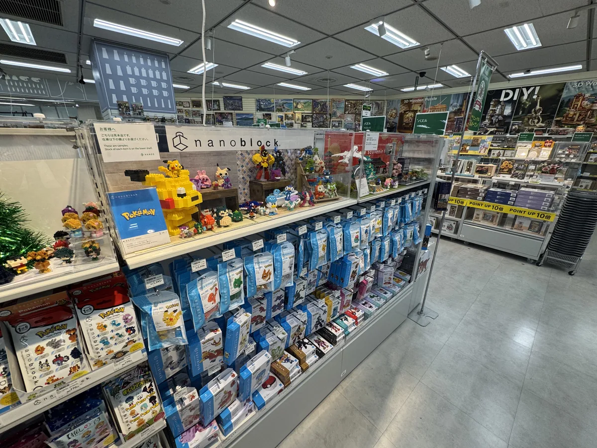Confezioni di piccoli giocattoli a mattoncini allineate sugli scaffali; in una vetrina trasparente in alto ci sono modelli montati con i mattoncini, tra cui dei Pokémon.