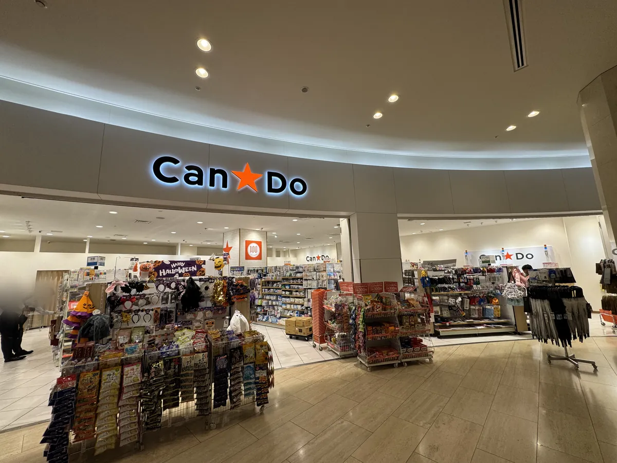Pasukan ng isang tindahan ng Can Do sa loob ng isang commercial facility. May karatula ng Can Do na may simbolong bituin sa puting pader, at may istante ng mga seasonal na produkto sa harap ng pasukan.