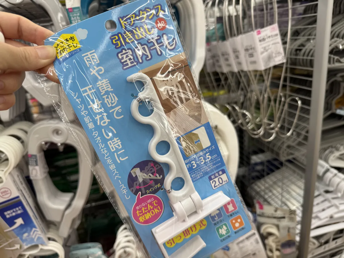 Hawak sa kamay ang pakete ng kawit ng DAISO na pansampay sa loob ng bahay. Sa asul na karton ay may paliwanag sa Japanese at larawang nagpapakita ng paggamit nito na nakasabit sa pinto.