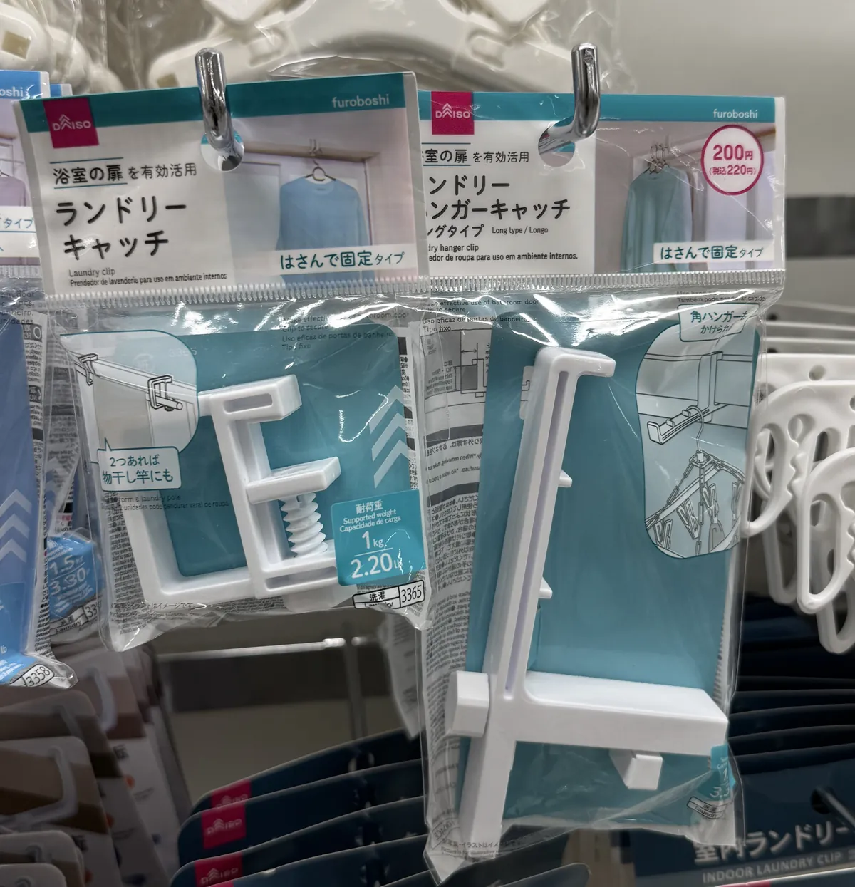 Dalawang pakete ng sipit ng damit (clothespin) ng DAISO. Sa kanang itaas ng paketeng nasa kanan ay may pulang bilog na markang "200円(税込220円)" (200 yen, 220 yen kasama ang buwis), at nakasulat sa Japanese at English ang pangalan ng produkto.