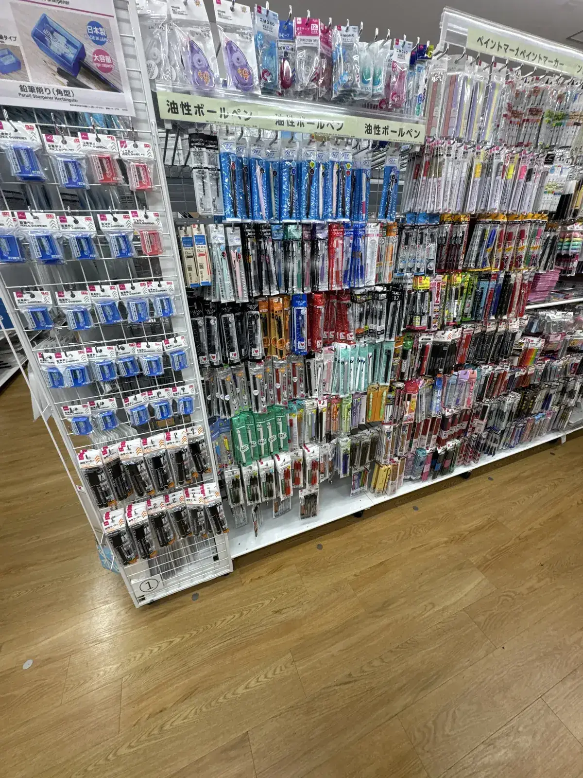 Seksyon ng stationery sa DAISO. May mga pantasa ng lapis sa kaliwa, maraming uri ng ballpen sa gitna at kanan, at karatulang "油性ボールペン" (oil-based ballpen) sa itaas ng istante.