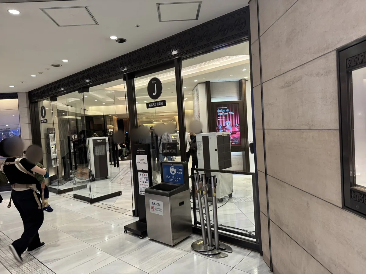 ทางเข้าหนึ่งของอาคารหลักของ Isetan Shinjuku เหนือประตูกระจกมีป้าย "Gate J 新宿三丁目駅側" (ฝั่งสถานี Shinjuku-sanchome) และข้างประตูมีประกาศเวลาเปิดทำการ (10:00am–8:00pm)