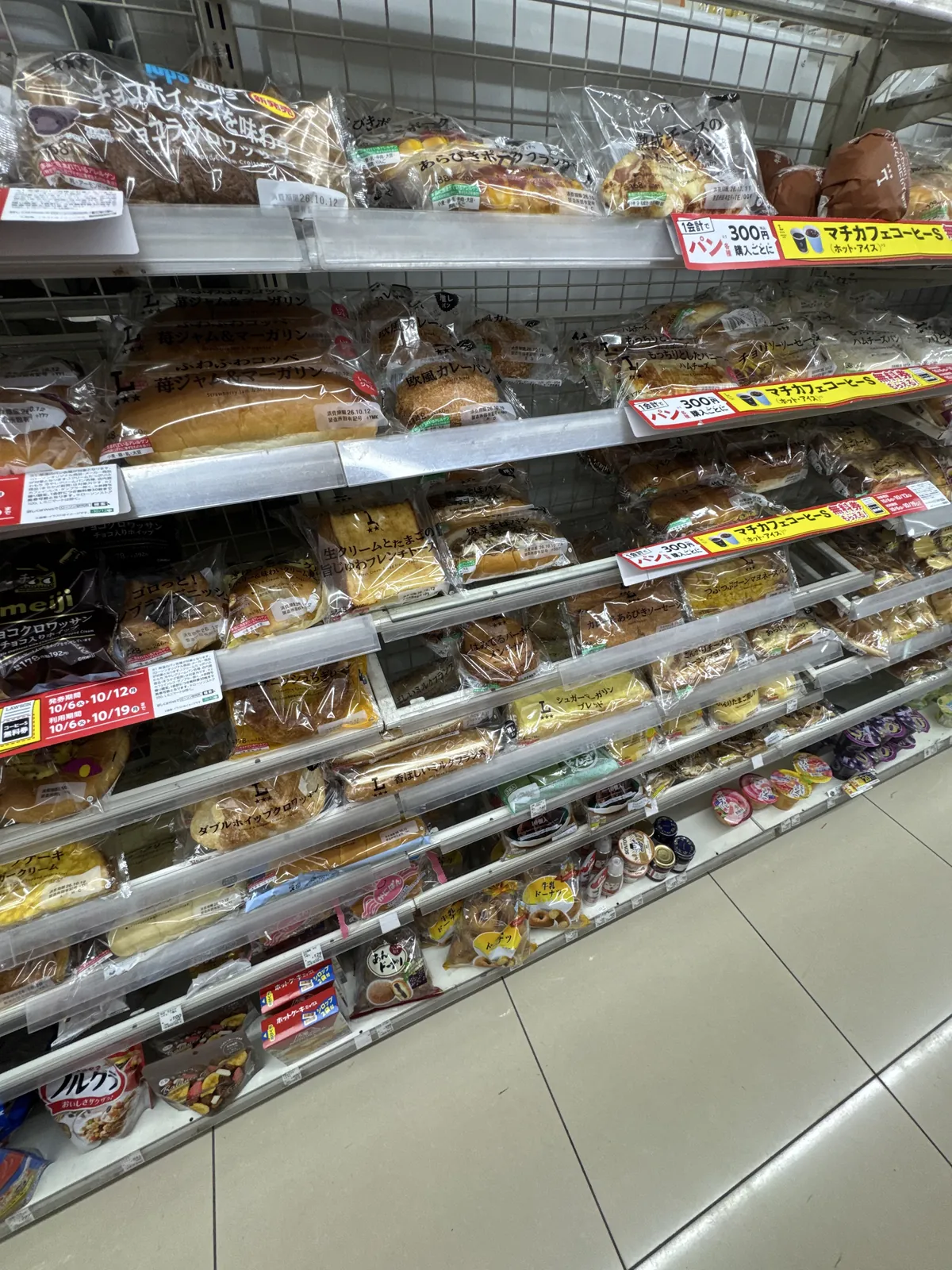 Rak roti di Lawson. Roti manis, roti berinti lauk dan kek gulung dalam beg disusun pada beberapa lapis rak, dan label rak memaparkan promosi yang memberikan kupon kopi.