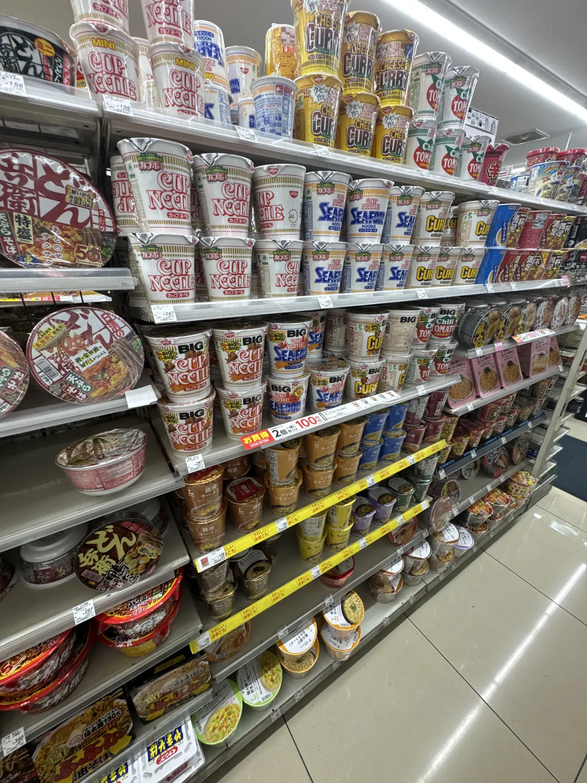 Rak mi cawan di Lawson. Mi cawan seperti Cup Noodle dan mi dalam bekas berbentuk mangkuk disusun pada beberapa lapis rak, dengan label harga.