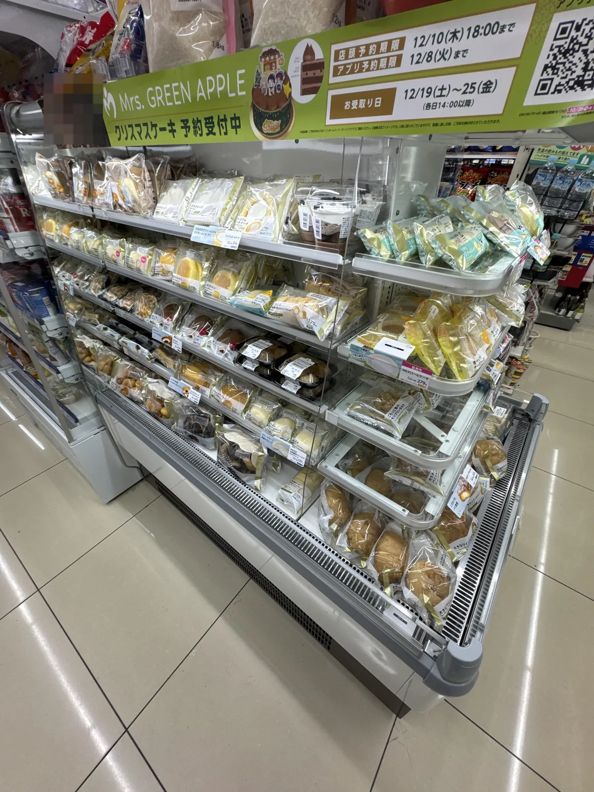 Rak sejuk pencuci mulut di Lawson. Kek dan puf krim dalam beg serta pencuci mulut dalam cawan disusun, dan di atasnya terdapat label tempahan kek Krismas. Wajah orang dalam gambar pada label dikaburkan dengan mozek.