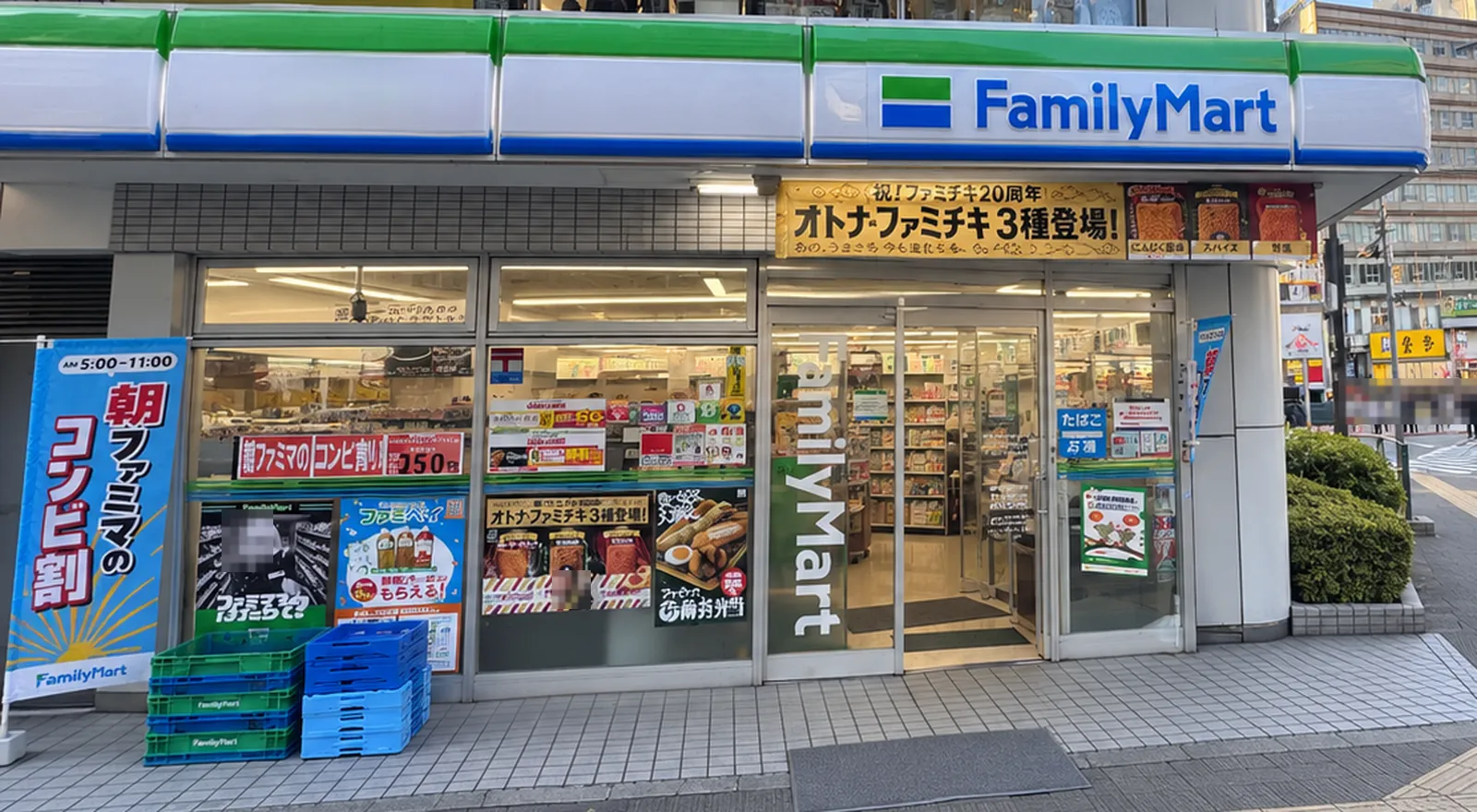 Bahagian depan kedai FamilyMart dilihat dari jalan. Di bawah jalur hijau dan biru serta papan tanda "FamilyMart" terdapat papan tanda ulang tahun ke-20 Fami-Chiki. Poster Fami-Chiki ditampal di tingkap, dan di sebelah kiri terdapat bendera menegak tentang diskaun waktu pagi. Wajah pada poster dan wajah pejalan kaki dikaburkan dengan mozek.