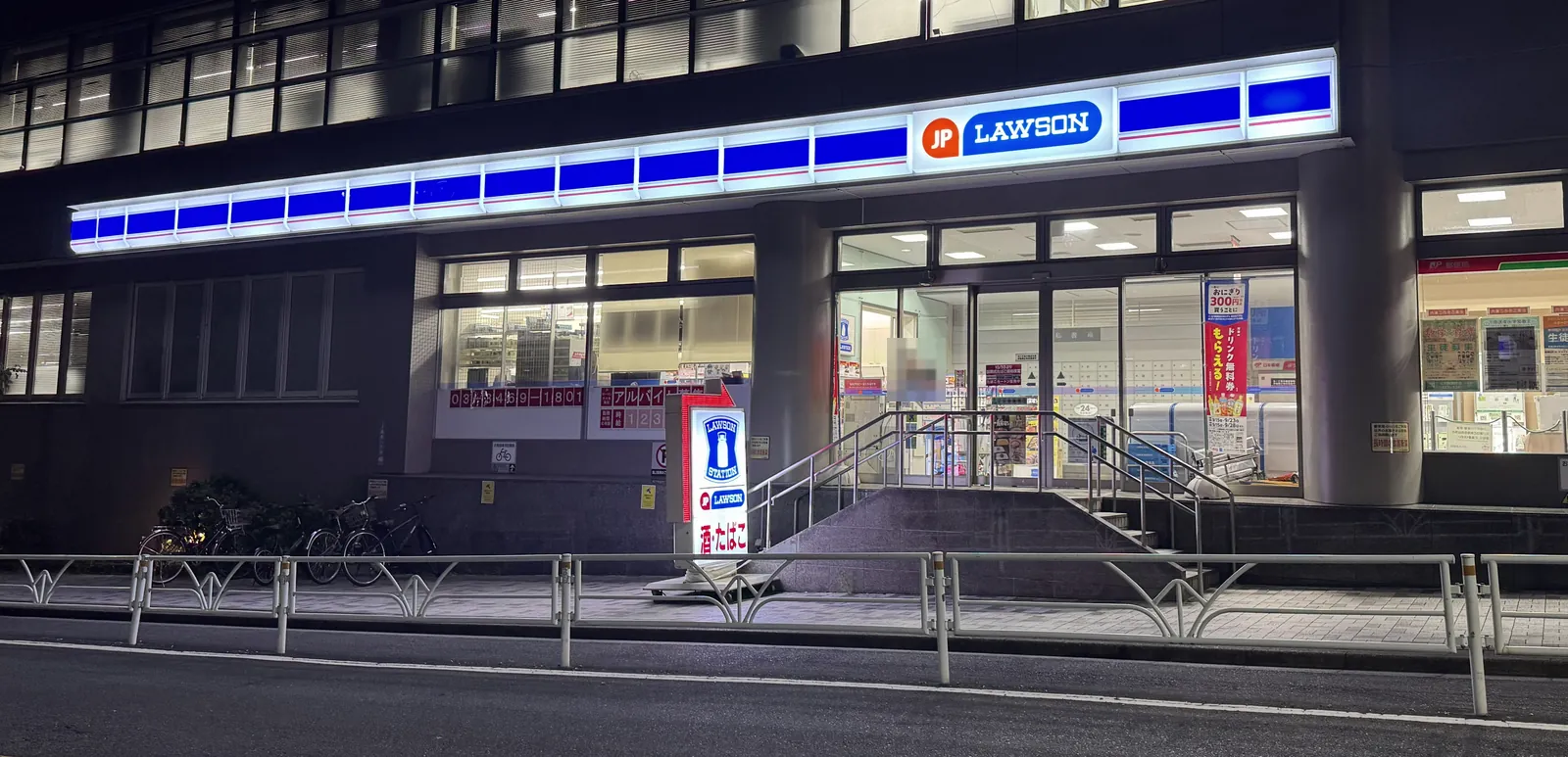 Bahagian depan kedai Lawson dilihat dari jalan pada waktu malam. Jalur biru dan papan tanda "LAWSON" bercahaya. Di depan pintu masuk terdapat anak tangga dan pemegang tangan, dan di sebelah kiri terdapat papan tanda "minuman beralkohol dan rokok". Wajah orang di dalam kedai dikaburkan dengan mozek.
