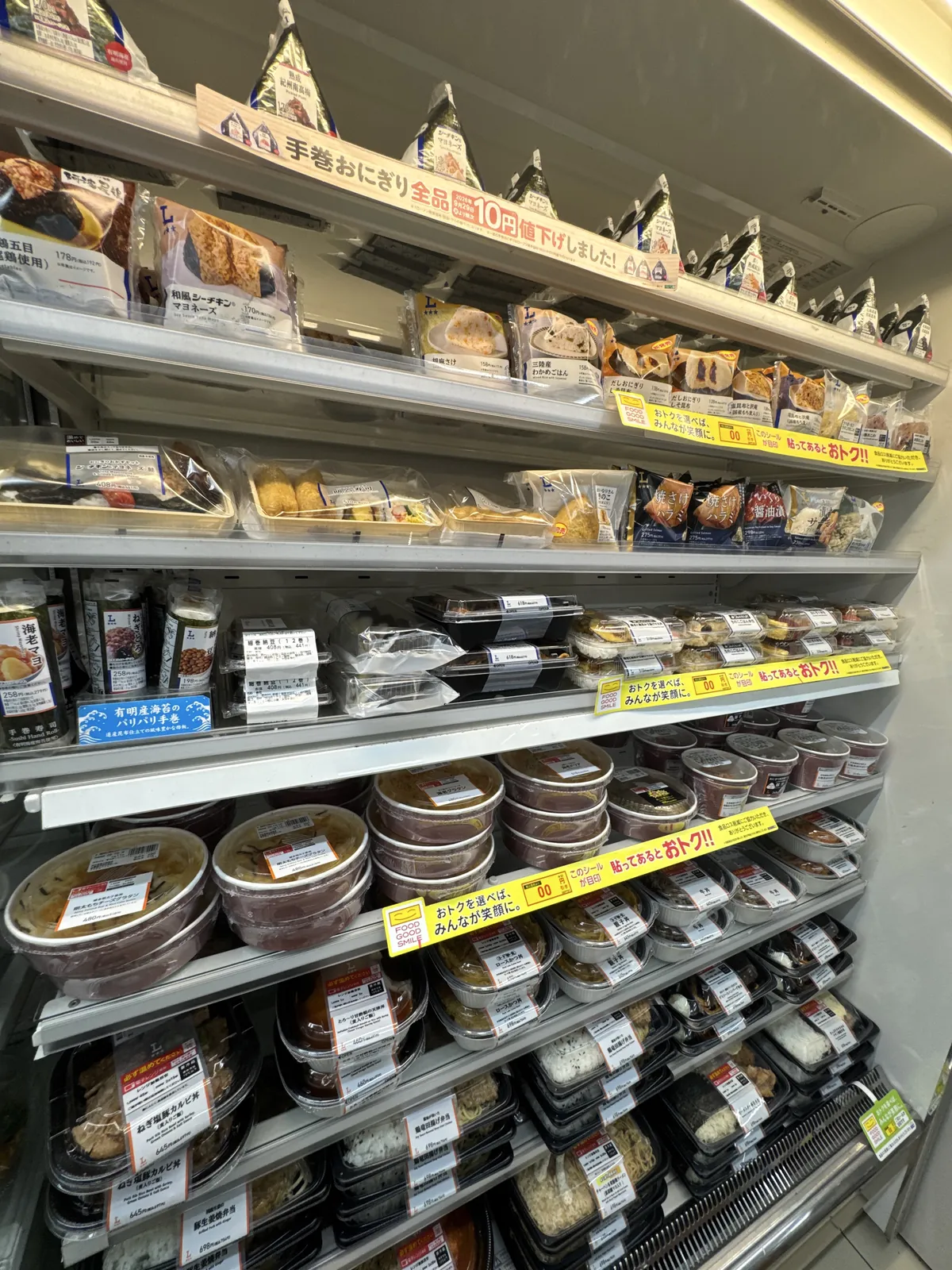 Rak sejuk di Lawson. Di rak atas terdapat onigiri segi tiga dan temaki (sushi gulung tangan), dan di beberapa lapis rak bawah terdapat donburi, bento dan mi, masing-masing dengan label harga.