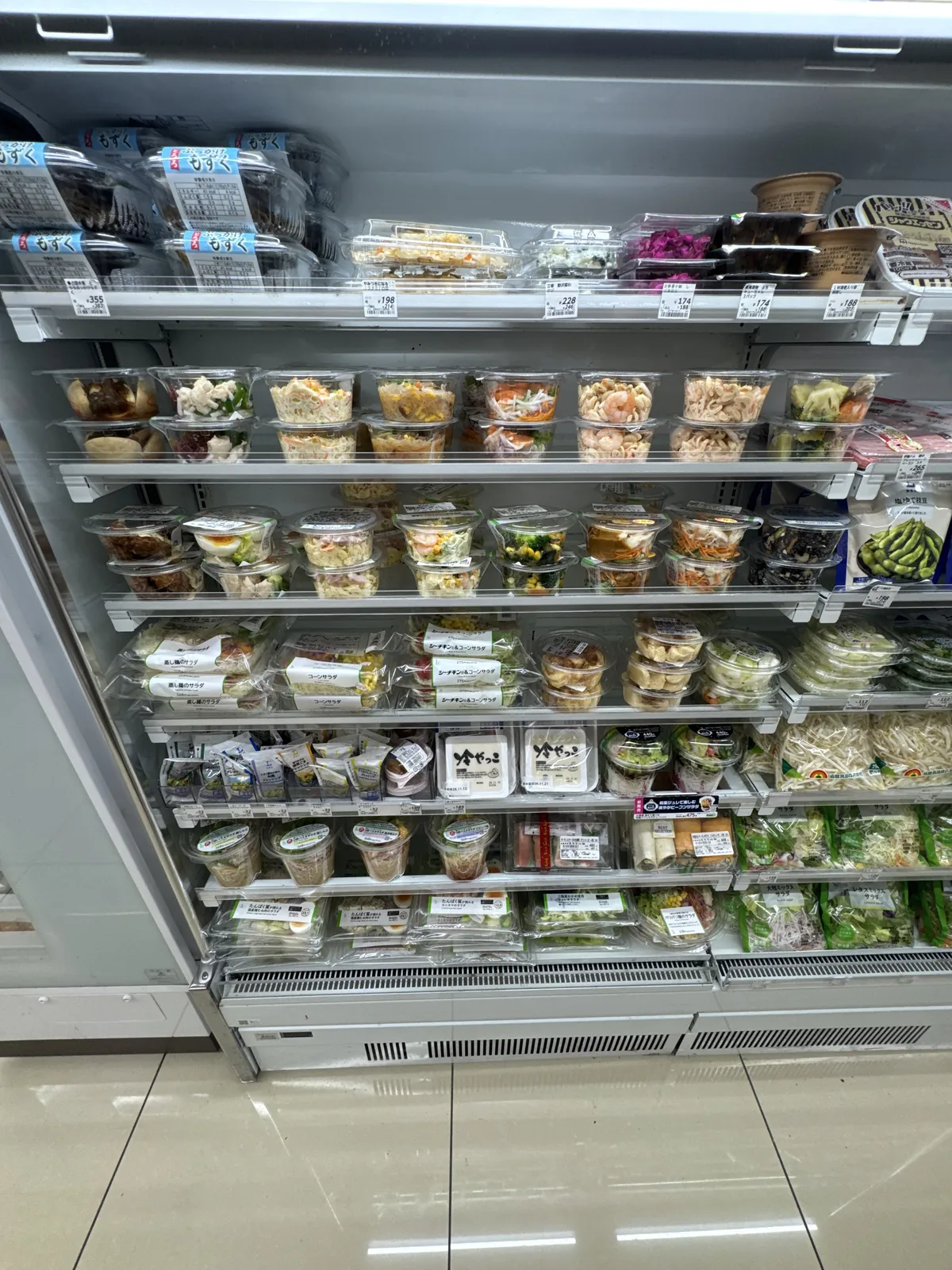 Rak sejuk di Lawson. Salad dan lauk siap masak dalam cawan, salad dalam beg dan tauhu disusun pada beberapa lapis rak, dengan label harga.