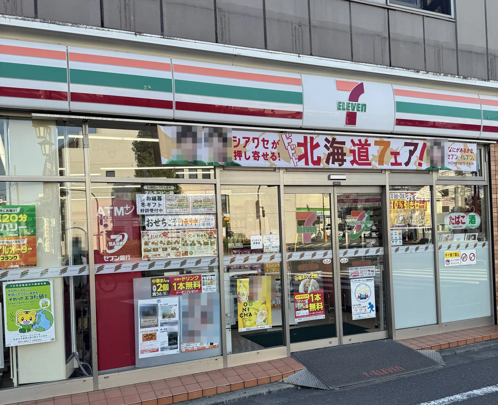 Bahagian depan kedai 7-Eleven dilihat dari jalan. Di bawah jalur hijau, oren dan merah serta papan tanda "7 ELEVEN" terdapat sepanduk "Pesta Hokkaido". Wajah orang pada sepanduk dan poster di pintu dikaburkan dengan mozek. Di sebelah kiri dinding kaca terdapat tanda ATM, dan di sebelah kanan tanda "rokok" dan "minuman beralkohol".