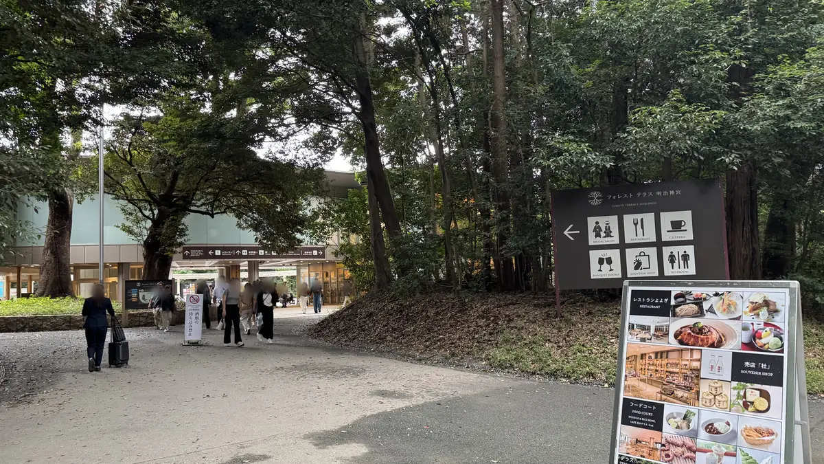 La strada davanti al Forest Terrace Meiji Jingu. A destra c'è un cartello direzionale con i disegni di ristorante, caffetteria self-service, negozio di souvenir e bagni. In fondo si vede un edificio con le pareti di vetro.