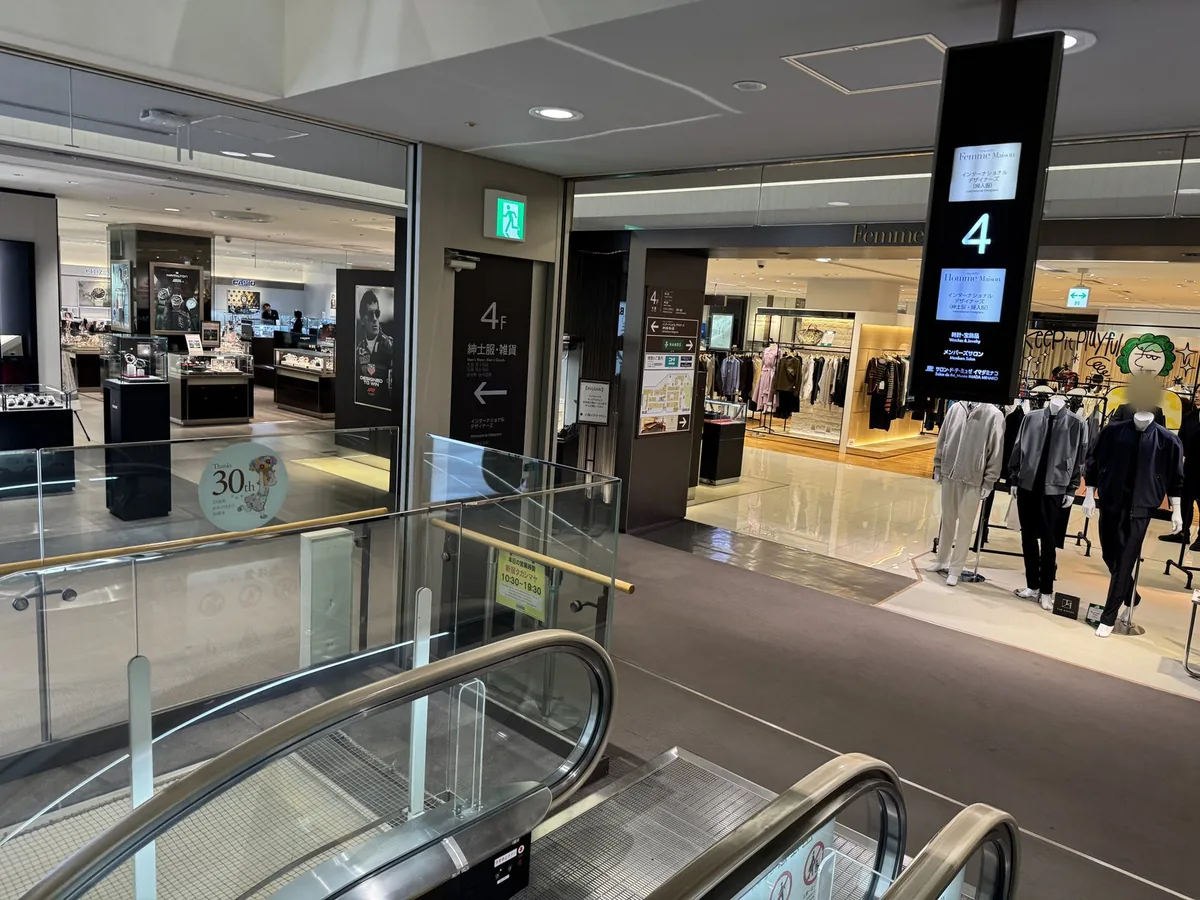Davanti alle scale mobili del 4F. Sul pannello informativo della colonna ci sono «4», «Femme Maison» e «Homme Maison», sulla parete «4F 紳士服・雑貨» (abbigliamento e accessori da uomo) e sul cartello in primo piano «本日の営業時間 新宿タカシマヤ 10:30〜19:30» (orari di oggi di Takashimaya Shinjuku: 10:30–19:30).
