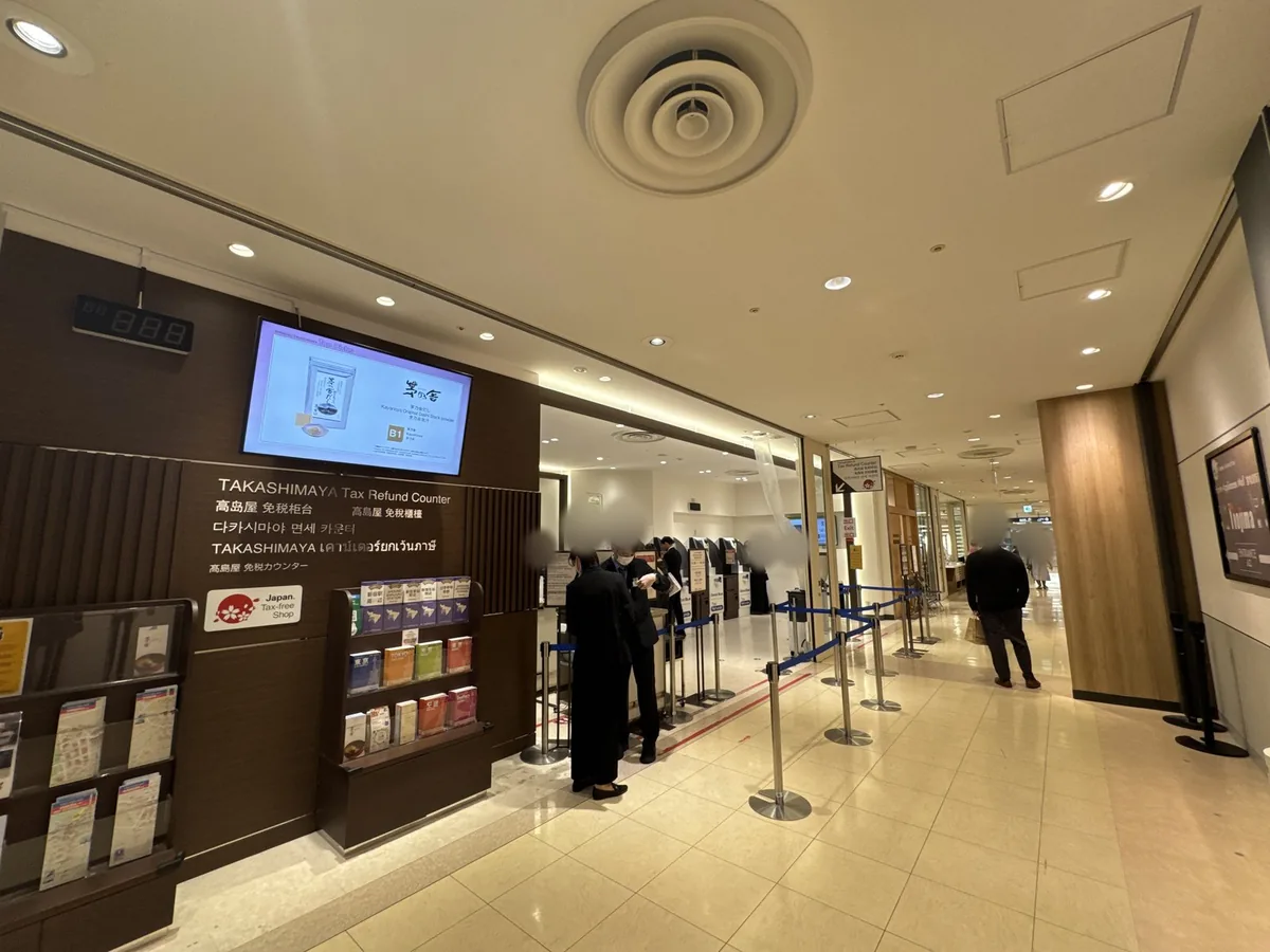 Ingresso dello sportello tax-free all'11F. Sulla parete di legno c'è la scritta «TAKASHIMAYA Tax Refund Counter» in inglese, cinese semplificato, cinese tradizionale, coreano, thailandese e giapponese; sotto ci sono il marchio «Japan. Tax-free Shop» e uno scaffale con opuscoli turistici nelle varie lingue.